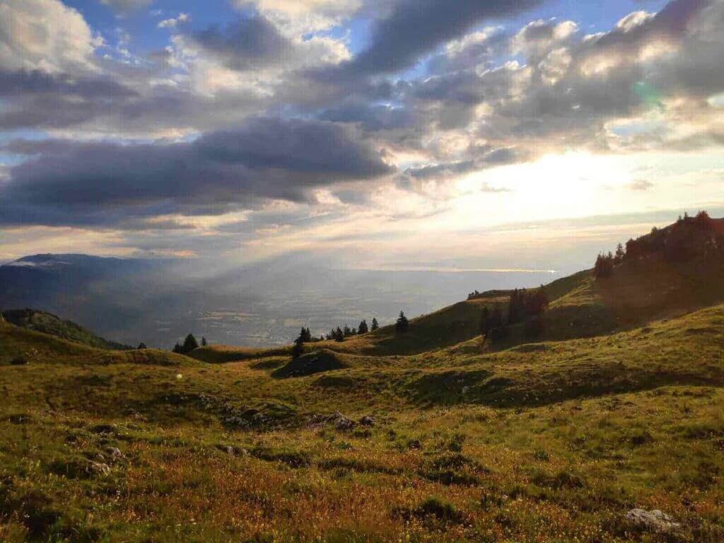 Rayons de soleil perçant les nuages au-dessus du bassin lémanique, vus depuis une crête herbeuse du Jura.