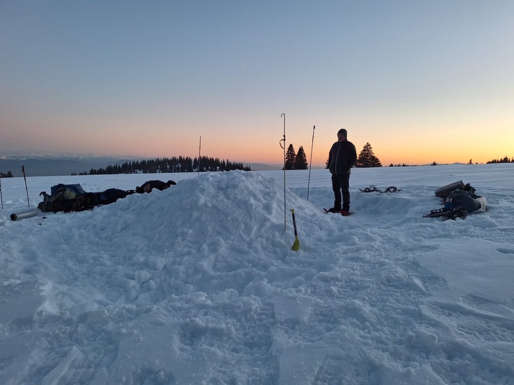 Campement de neige au coucher du soleil sur un plateau du Jura, une personne en raquettes debout près d'un abri en construction.